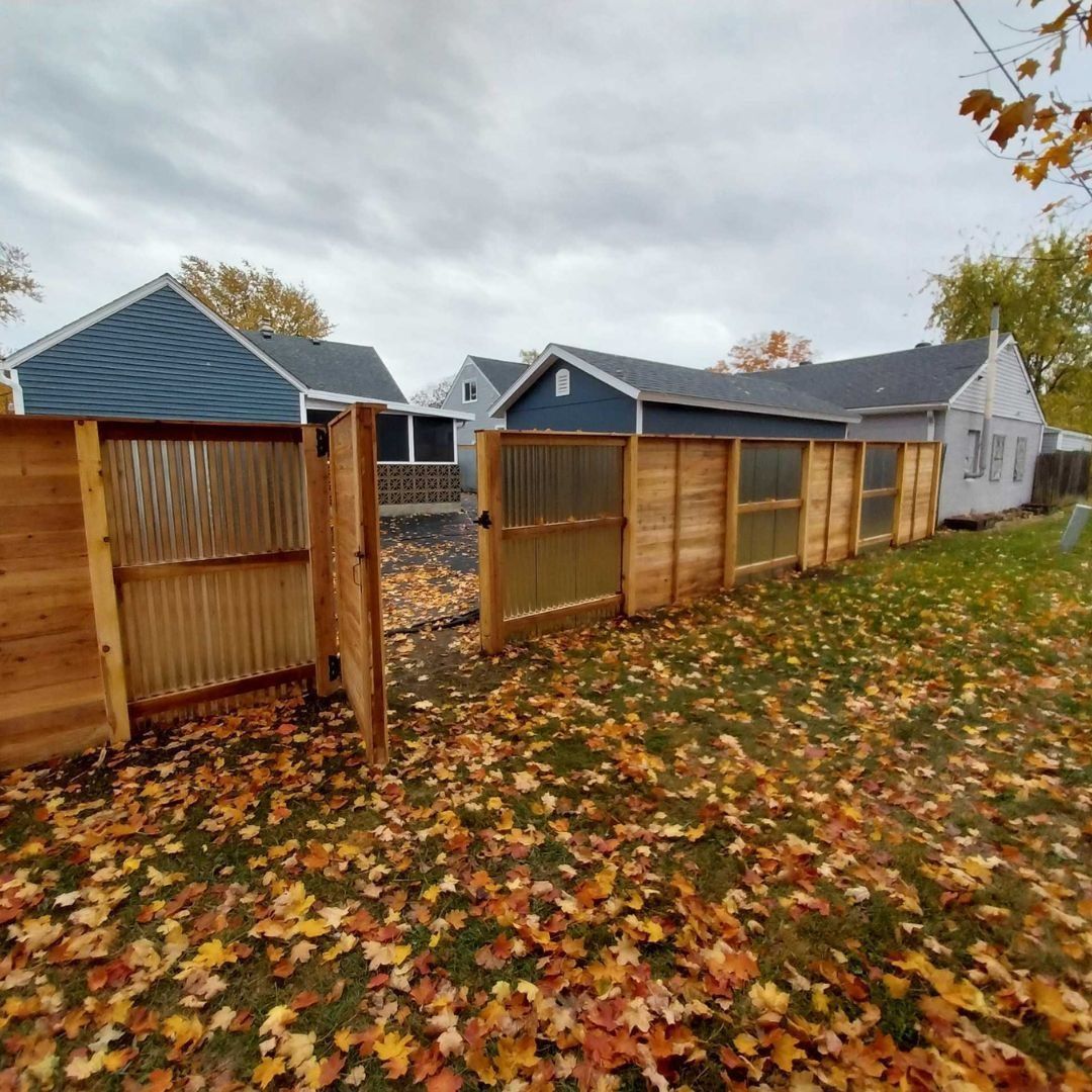 Wooden fence with gates in a yard covered in fallen leaves, houses in background under a cloudy sky.