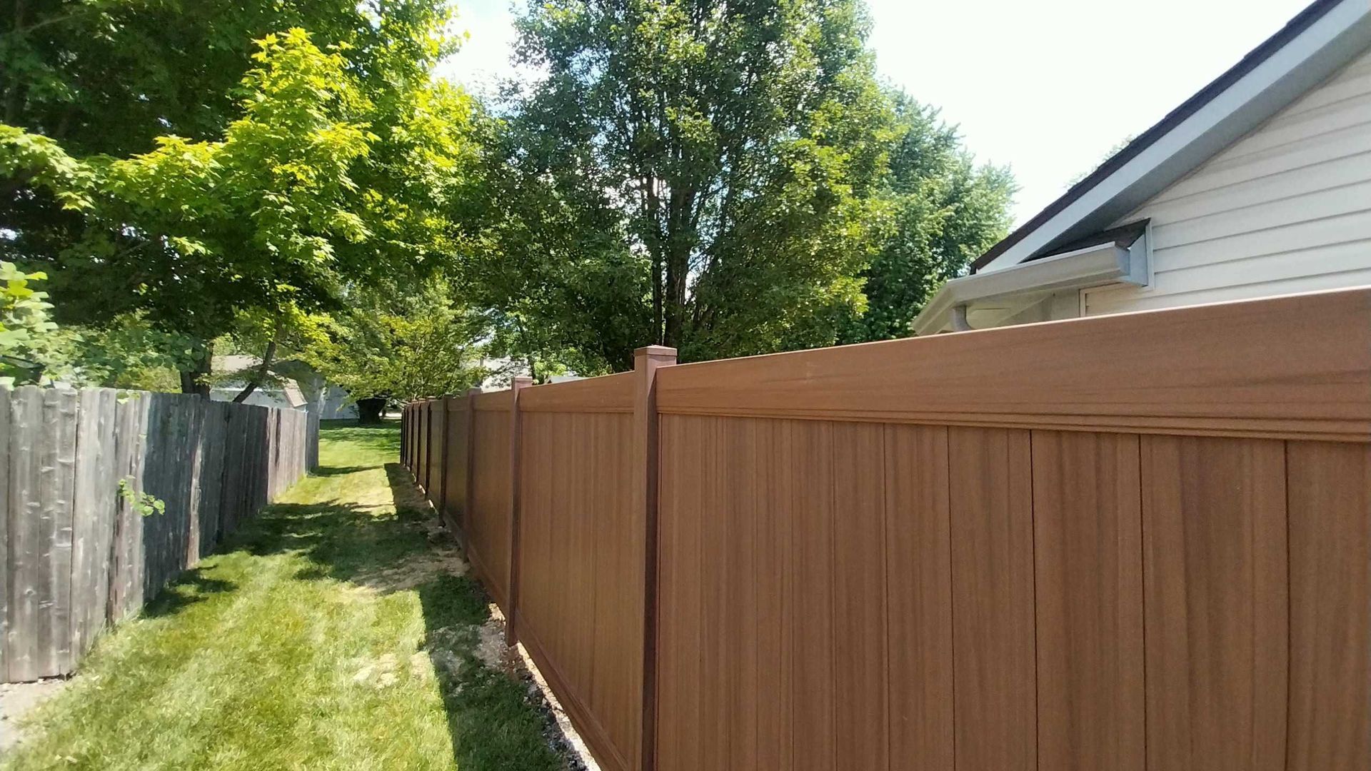 Brown wooden fence alongside a grassy area, trees, and a house with light siding.