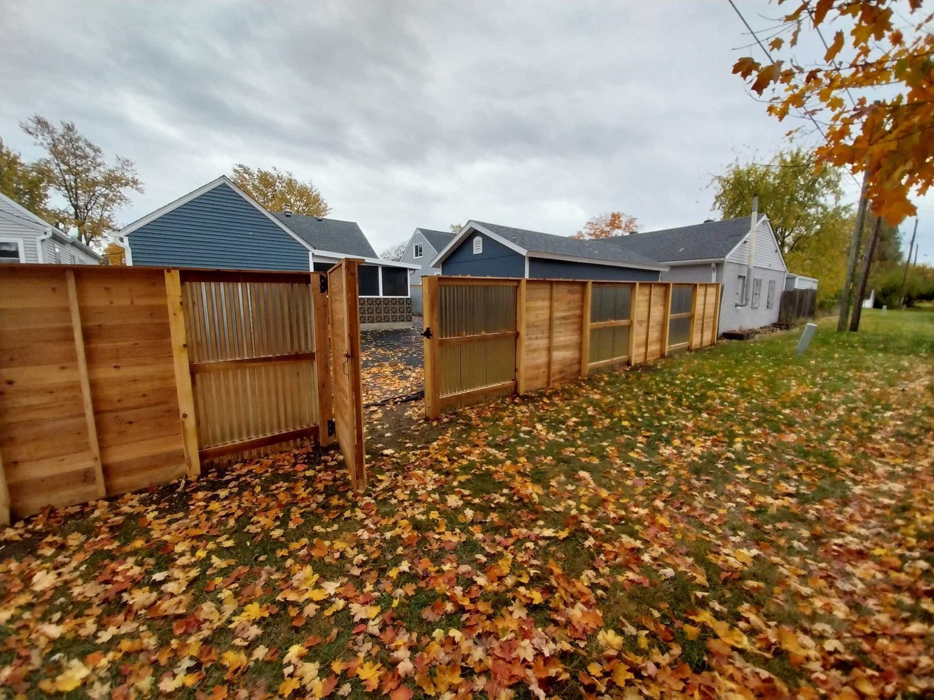 Wooden fence with corrugated panels surrounds a yard covered in fallen autumn leaves; houses in the background.