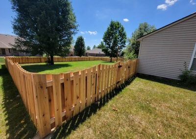 Wooden fence encloses a green lawn with trees and a light-colored house under a blue sky.