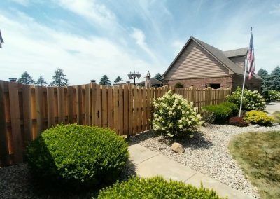 Wooden fence alongside a house with green bushes, gravel path, and American flag.