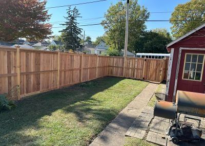 Wooden fence encloses a backyard with green grass and a small red building on a sunny day.