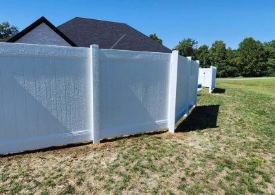 White vinyl fence in a grassy yard, partially obscuring a house with a dark roof.