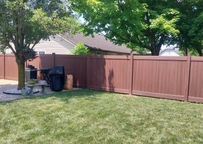 Brown wooden fence enclosing a green lawn, with a grill and trees visible.