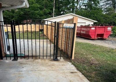 Black metal fence with wooden panel, red dumpster in background.