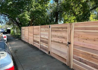Wooden fence with a gate along a sidewalk, trees overhead, car parked on the street.