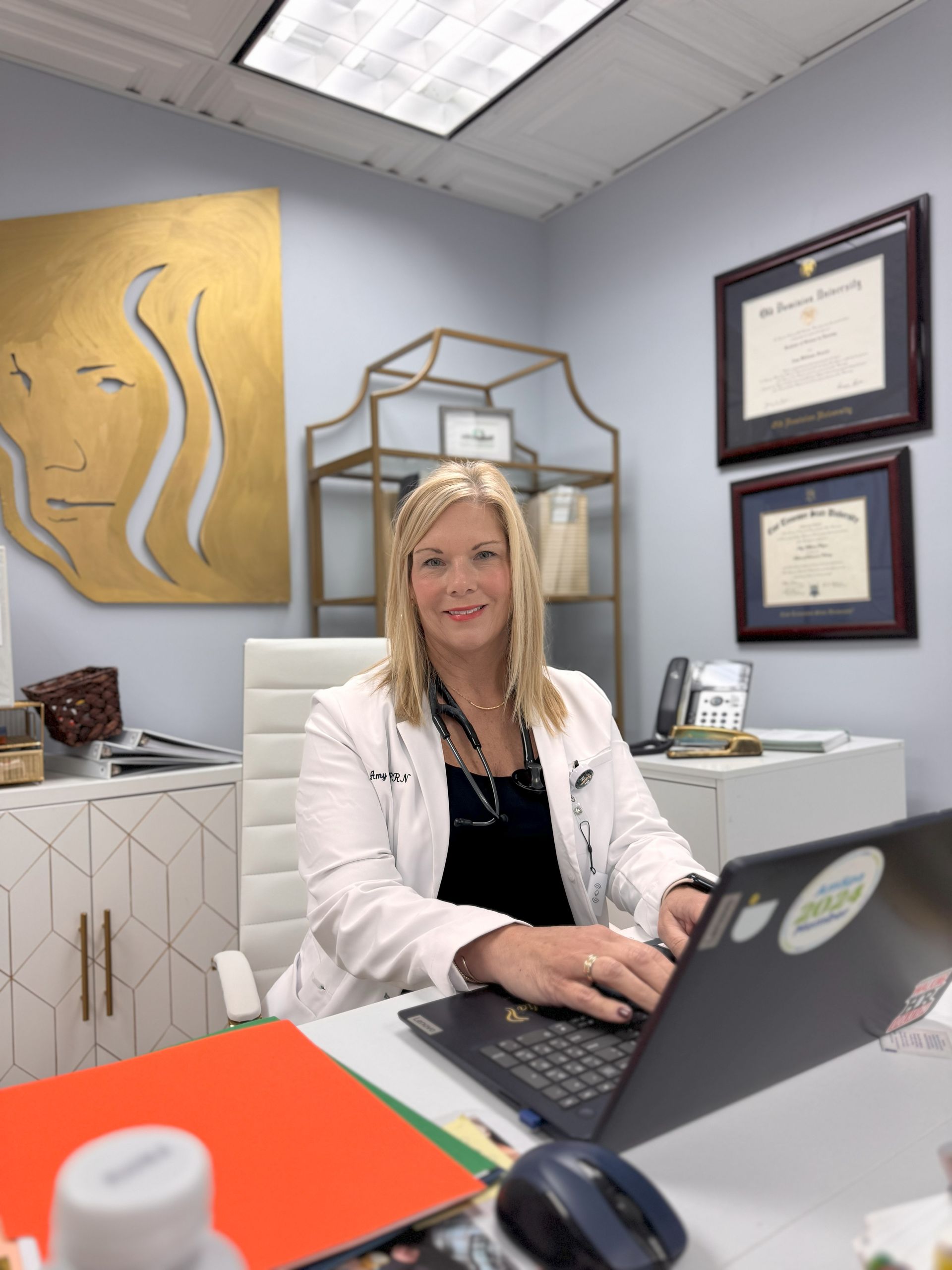 Woman in white coat at desk, typing on laptop in office. Gold face art on wall.