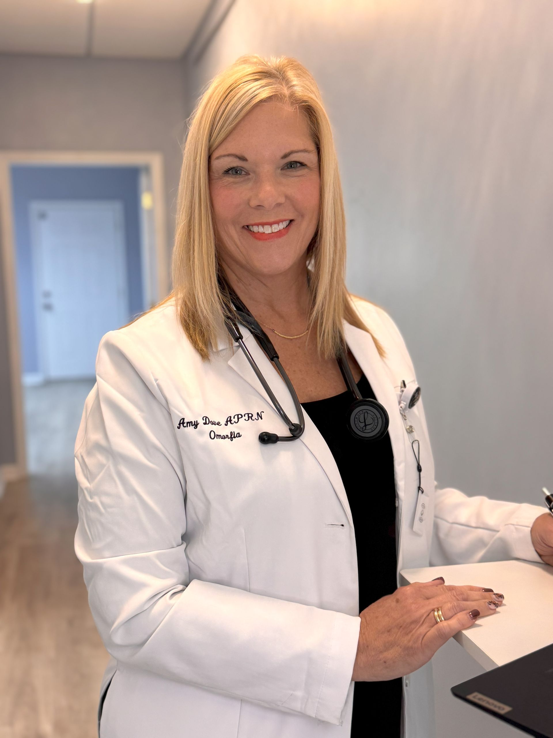 Woman in white lab coat with stethoscope, smiles while standing in a medical office doorway.