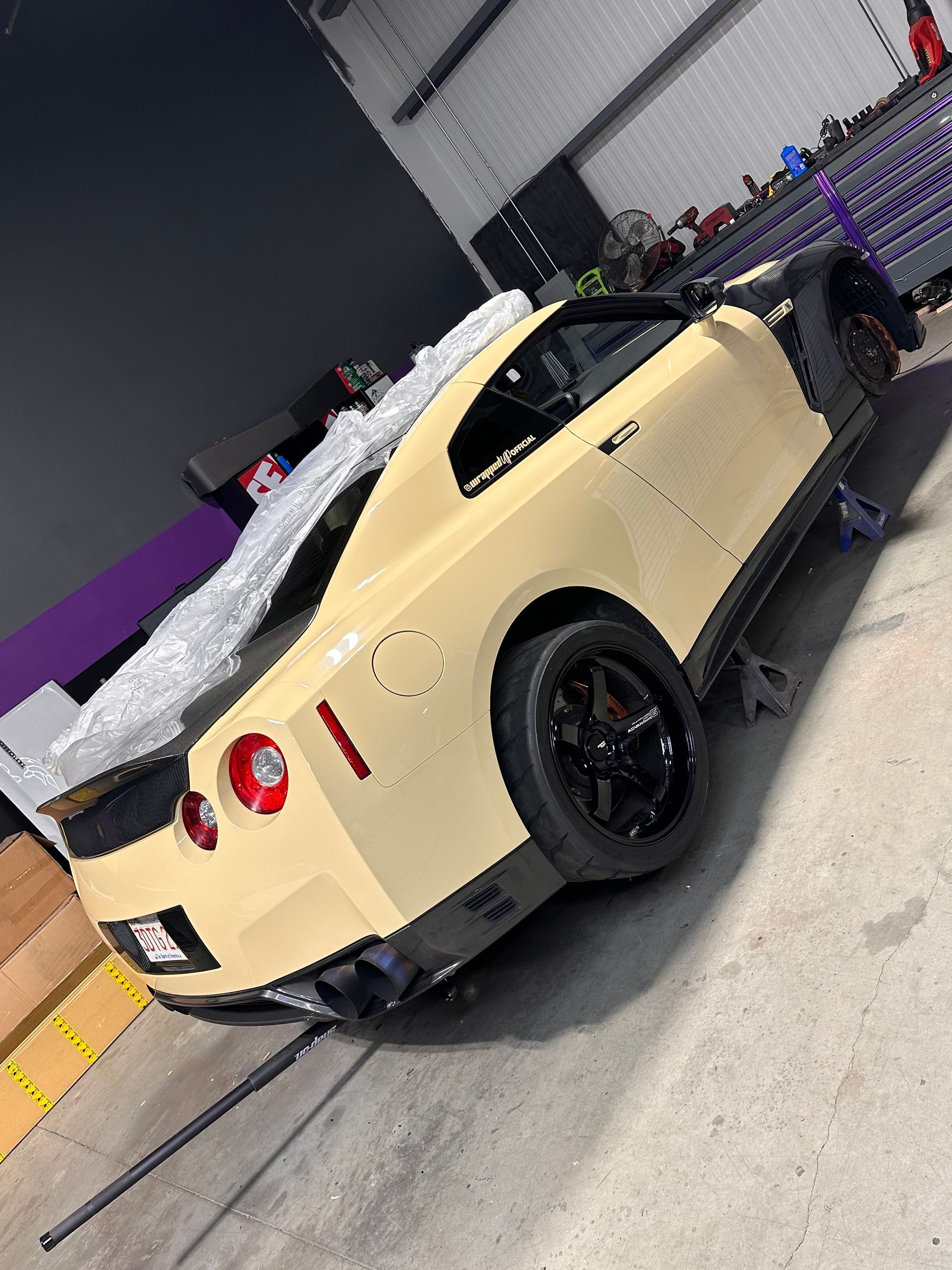 Tan Nissan GT-R sports car in a garage, on jack stands, black wheels, red tail lights, and black spoiler.