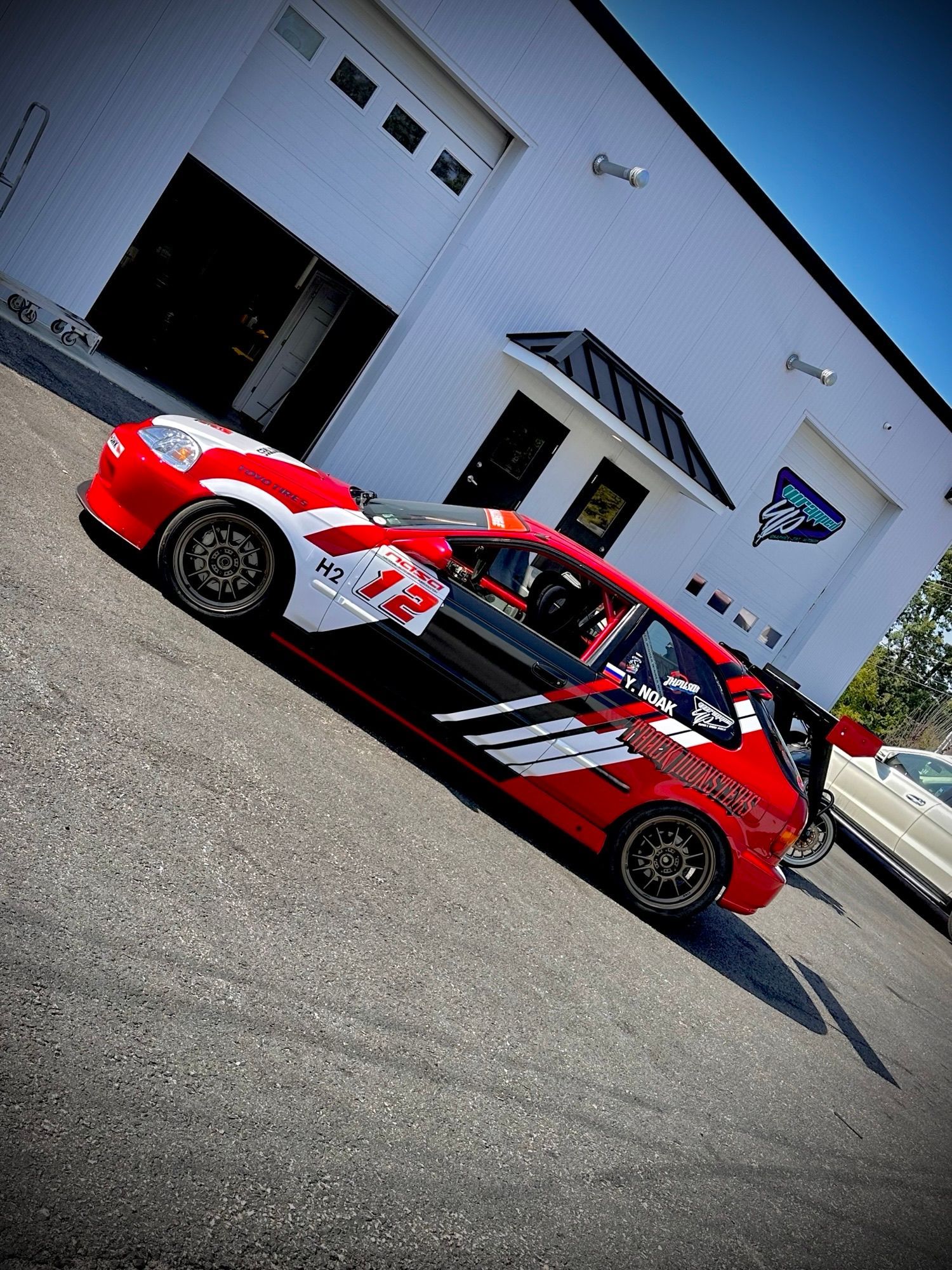 Red and white race car with number 12 parked outside a white building on a sunny day.