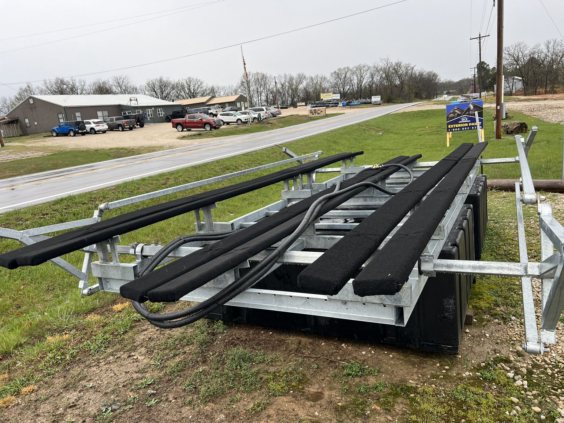 Boat trailer on a grassy area beside a road, gray metal frame with black padded bunks, overcast day.