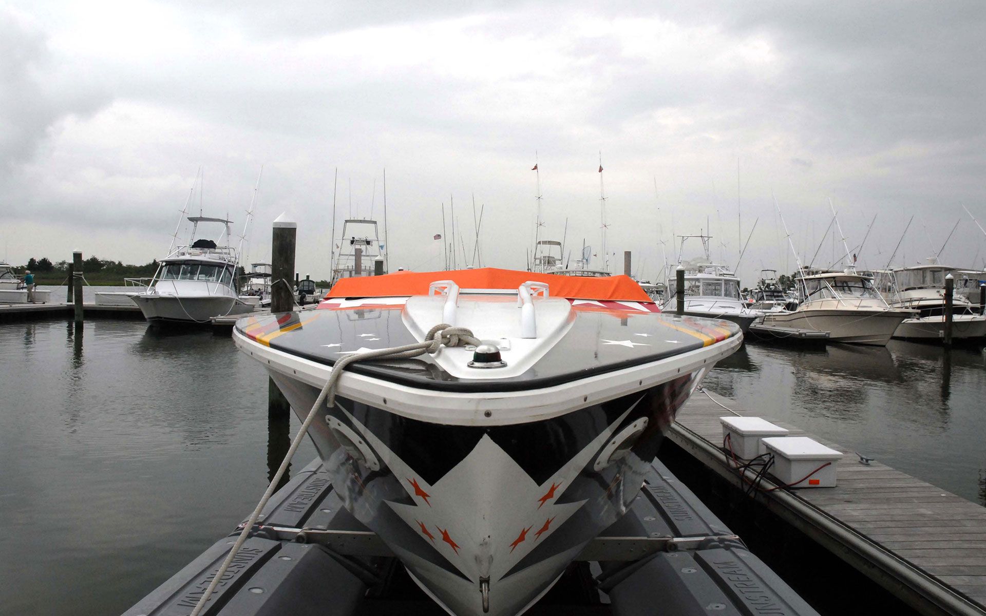 Speedboat docked at pier, facing camera, with other boats in background under overcast sky.