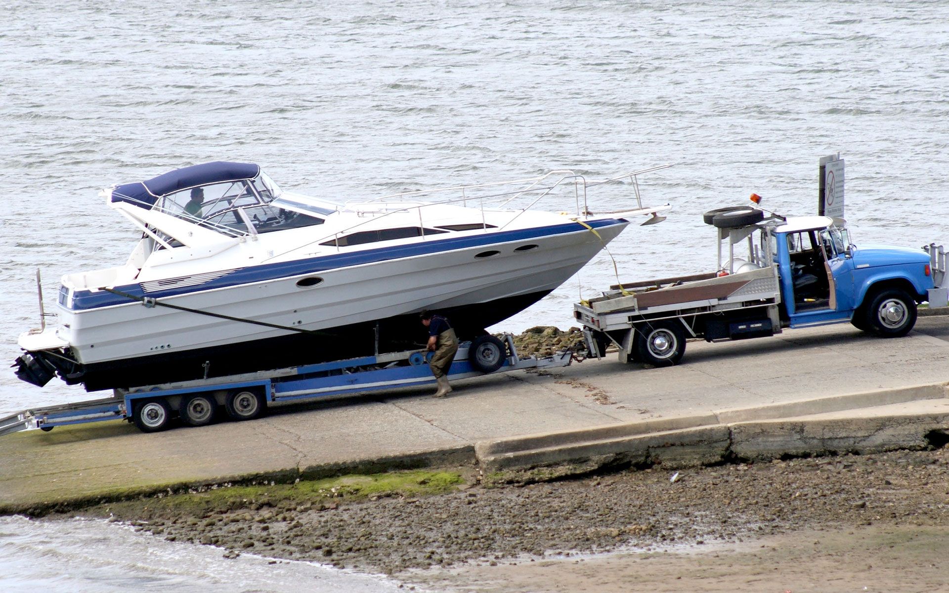A boat on a trailer being launched into water by a blue tow truck on a concrete ramp.