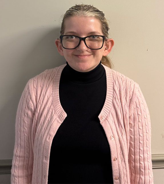 Woman wearing glasses, pink cardigan, and black turtleneck smiles in front of a neutral wall.