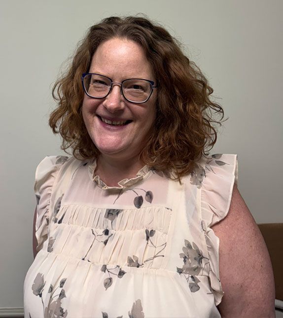 Woman smiling, wearing glasses and floral print top. Beige wall in background.