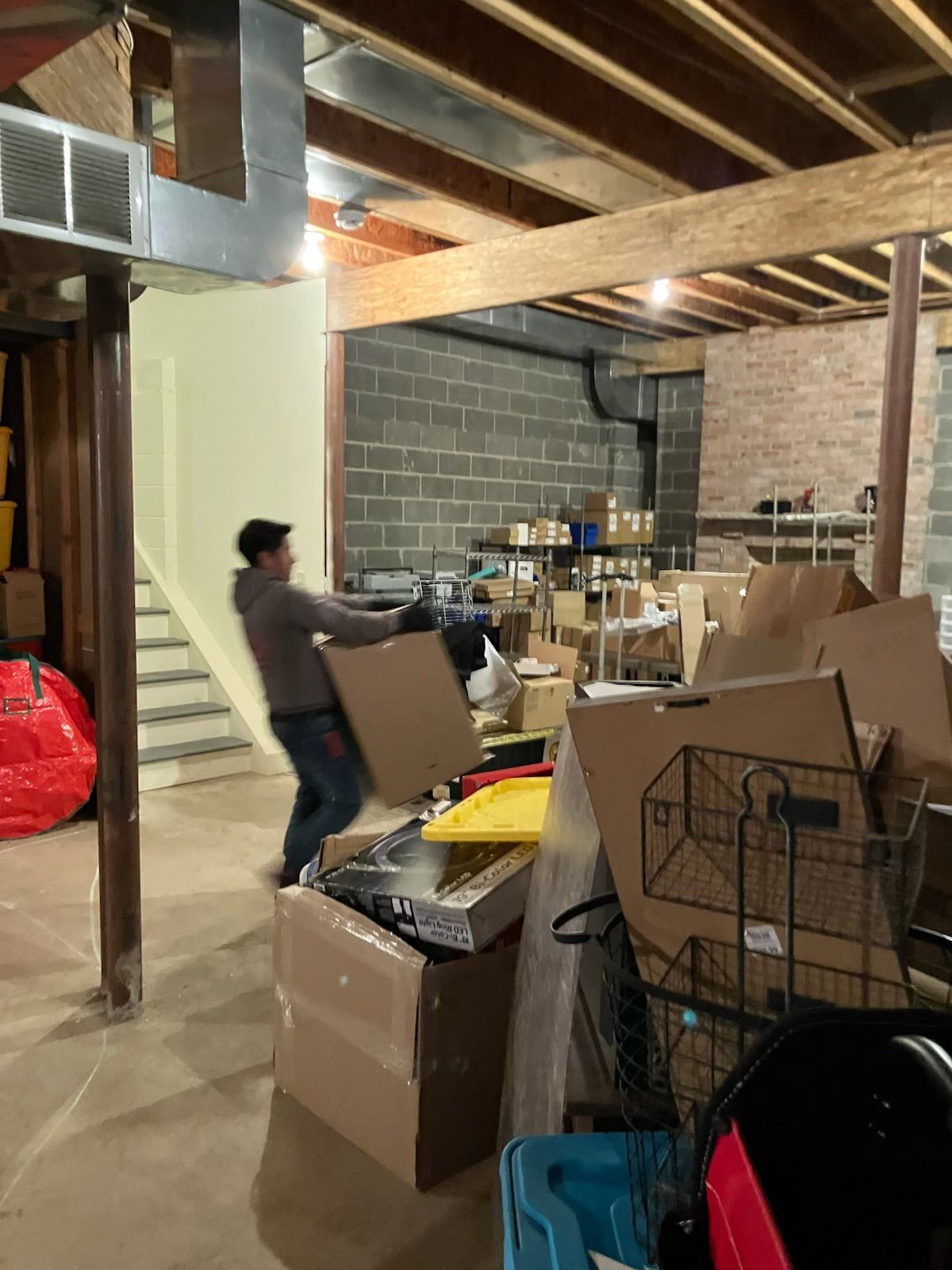 A man is carrying a cardboard box in a basement filled with boxes.