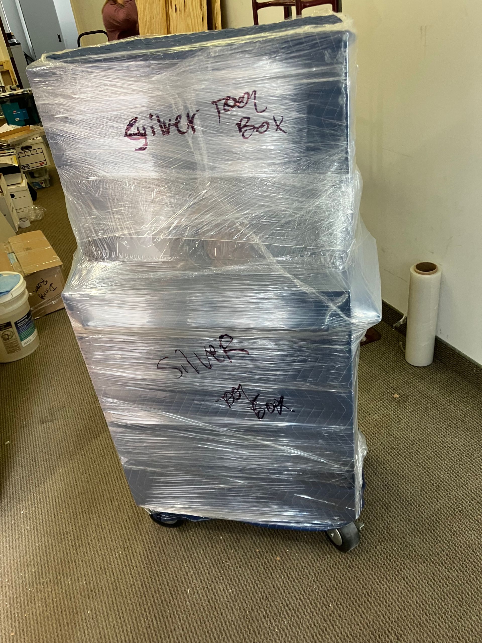 A metal cabinet is wrapped in plastic wrap and sitting on a carpet.