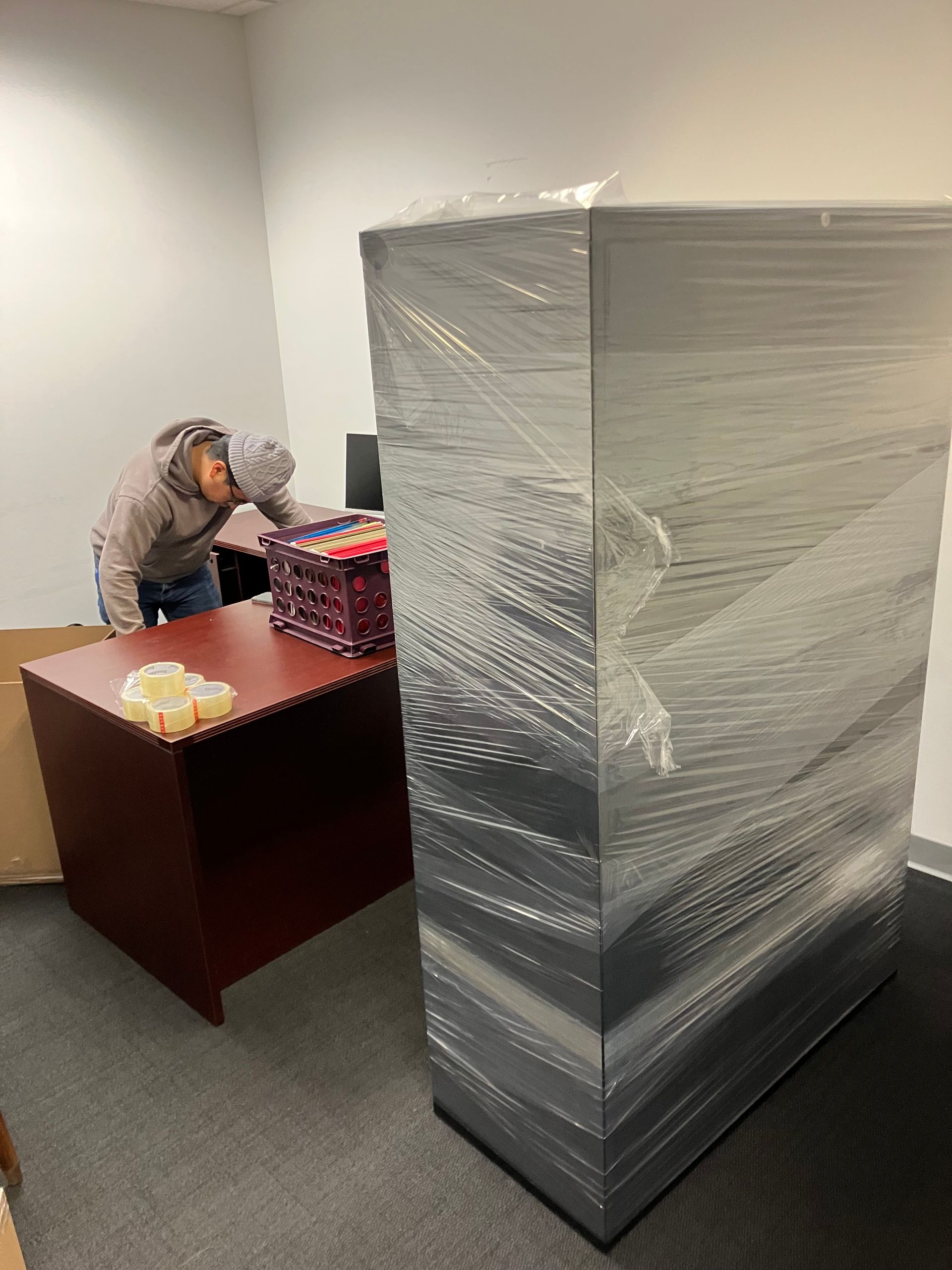 A man is wrapping a cabinet in plastic wrap in an office.