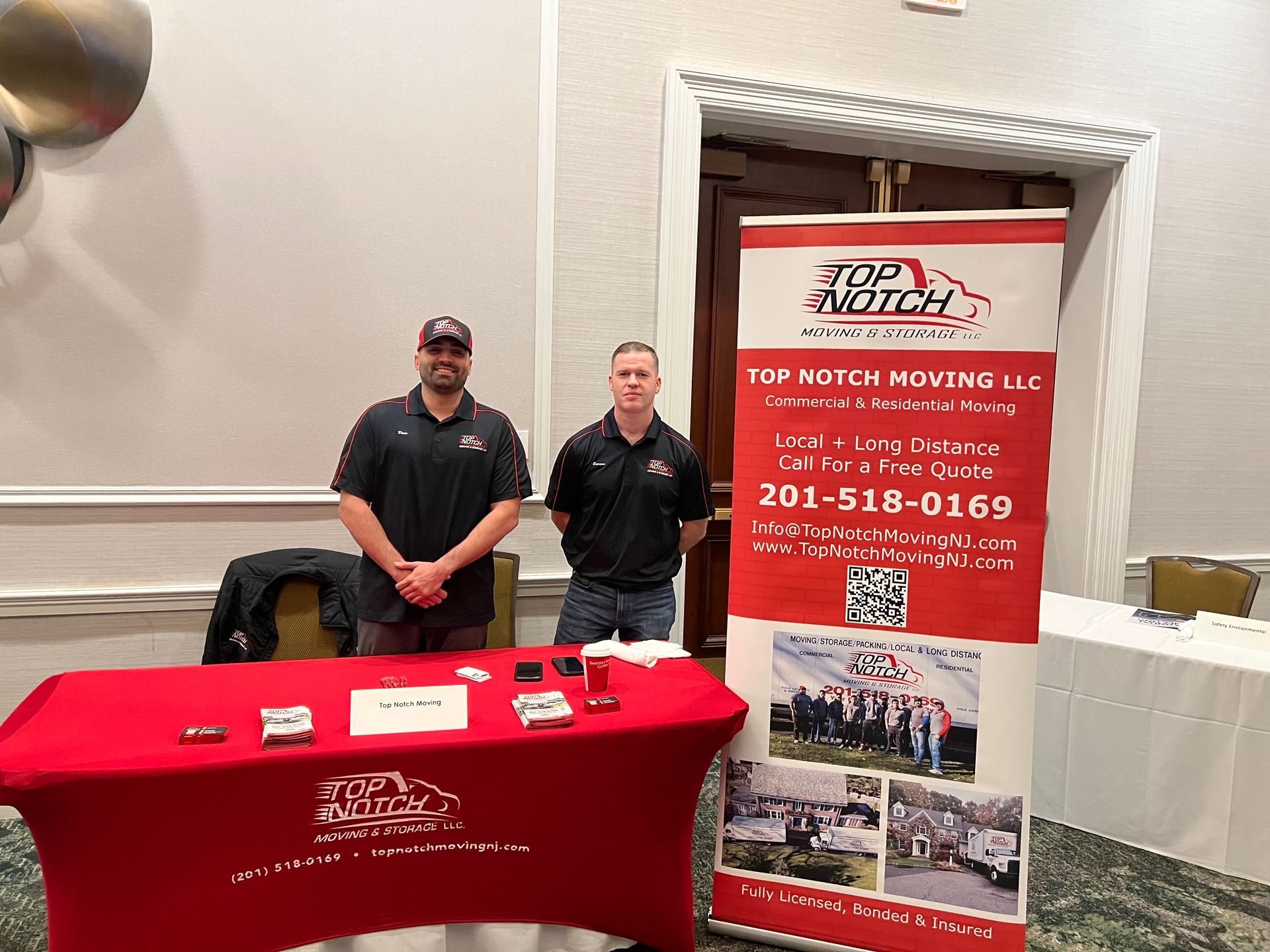 Two men are standing in front of a table with a sign on it.