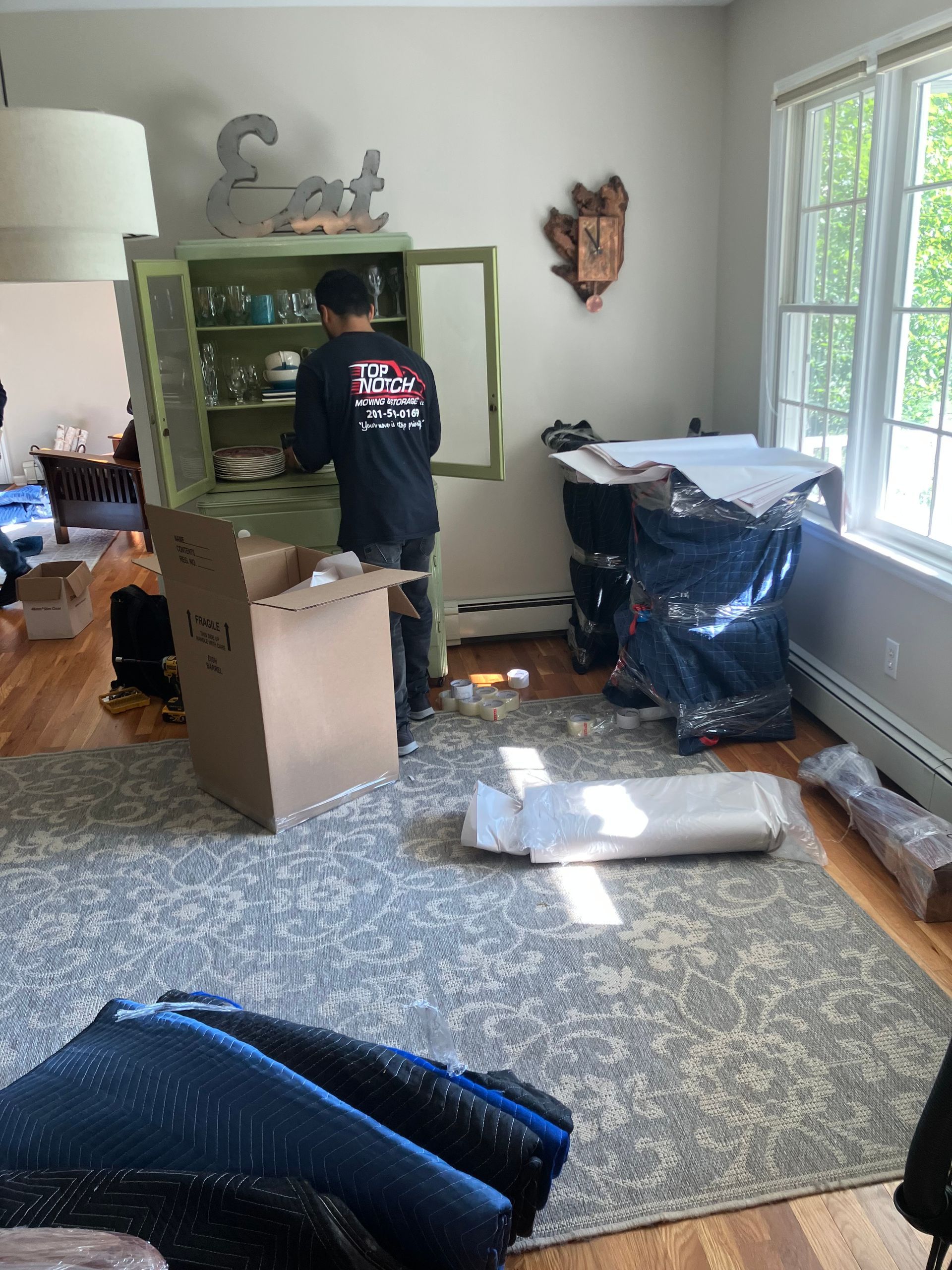 A person packing dishes in a green cabinet inside a living room with boxes, a rug, and a window.