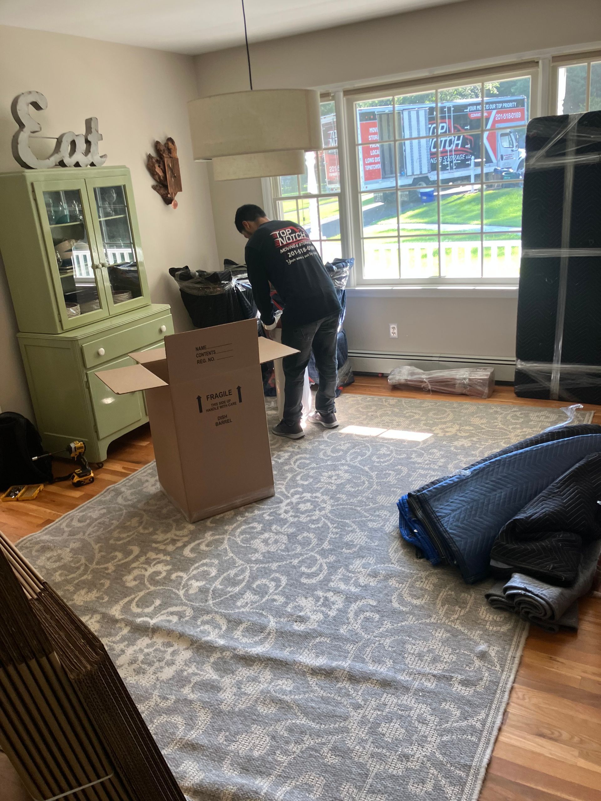 A person packing boxes in a room with a light green cabinet, a window, and wrapped furniture.