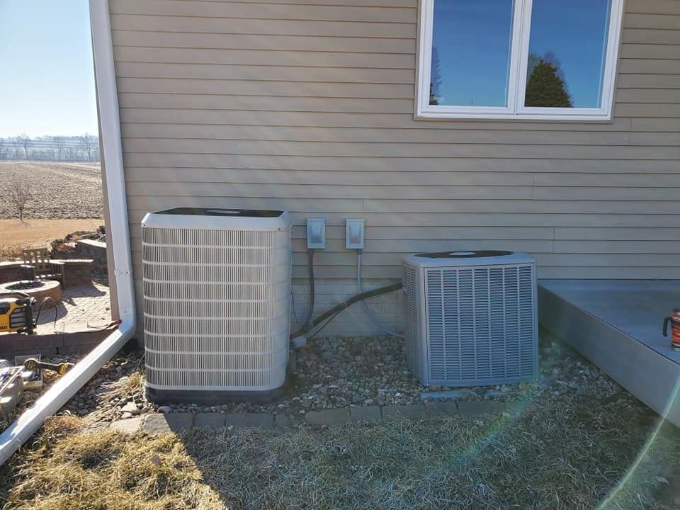 Two AC units next to a house with beige siding, gravel ground. Clear sky.