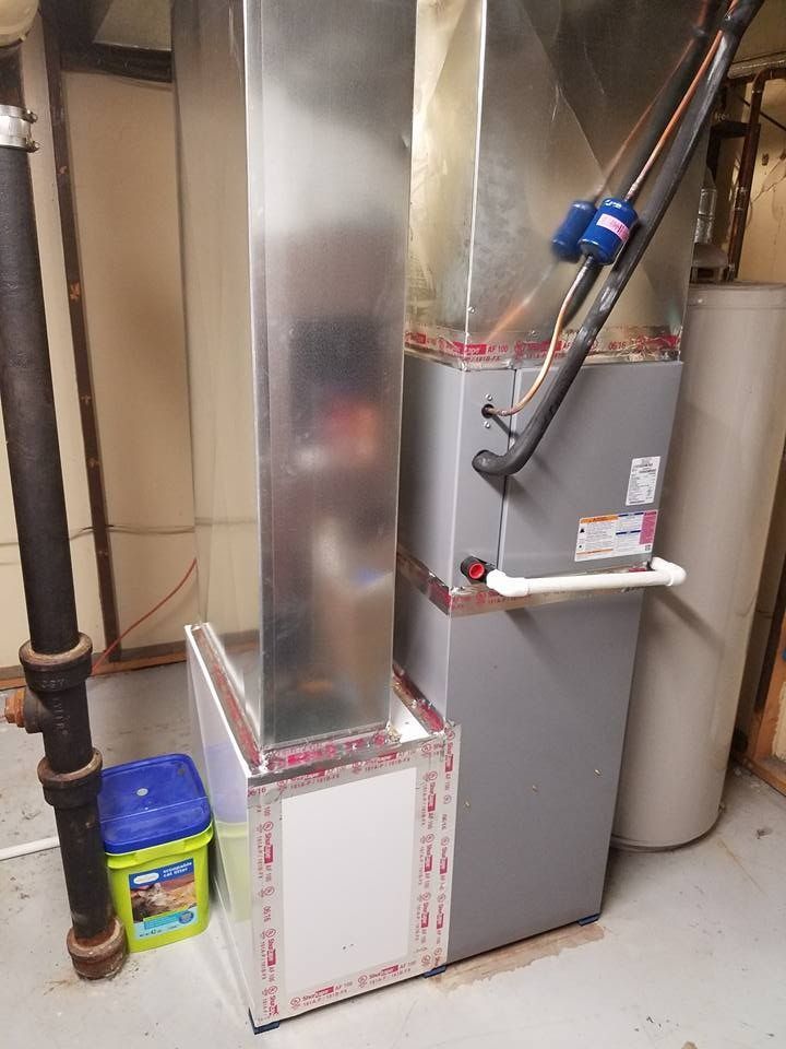Furnace and ductwork in a basement. Silver metal ducts, grey furnace, water heater, and a blue container.