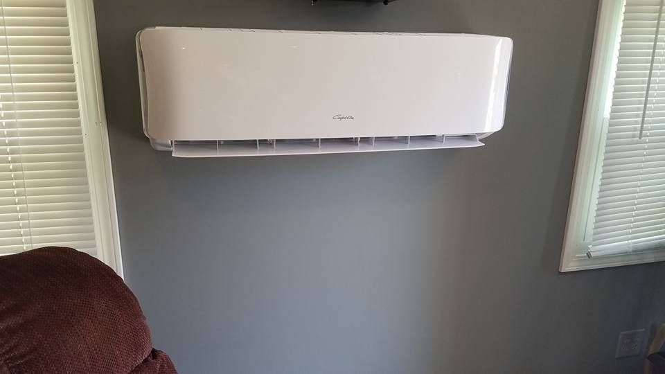 White ductless mini-split air conditioner mounted on a gray wall between two windows with blinds.