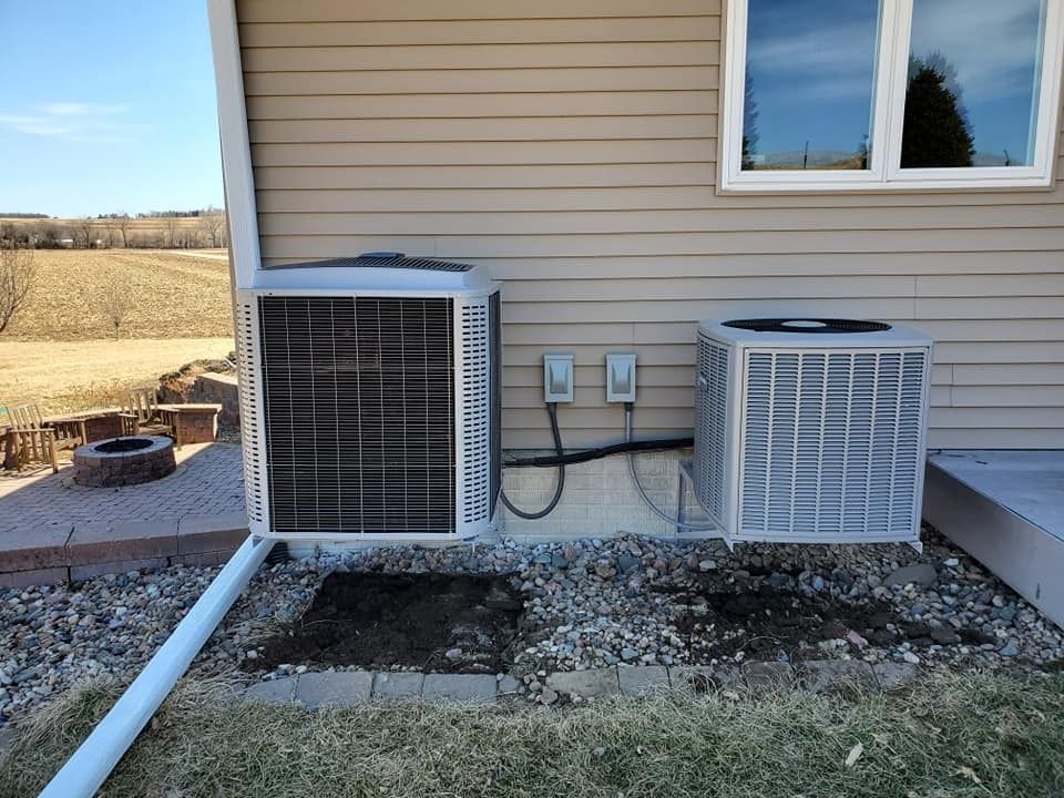 Two air conditioning units beside a house on a gravel bed; blue sky and field in the background.
