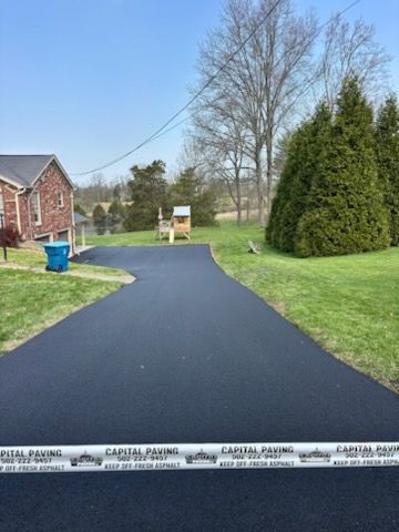 A freshly paved black asphalt driveway stretches toward a brick house, blocked by caution tape from Capital Paving.