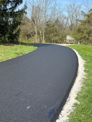 A freshly paved, smooth black asphalt driveway curves through a grassy, tree-lined landscape with a gravel border.