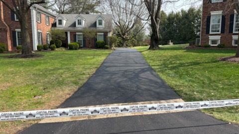 A freshly paved asphalt driveway sits between two brick homes, blocked by caution tape marked 