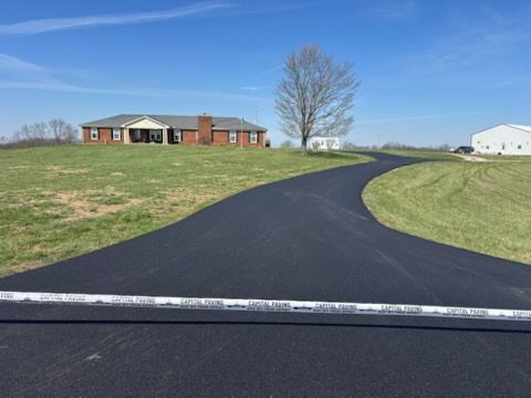 A newly paved asphalt driveway leads toward a brick house on a grassy lot under a clear blue sky.