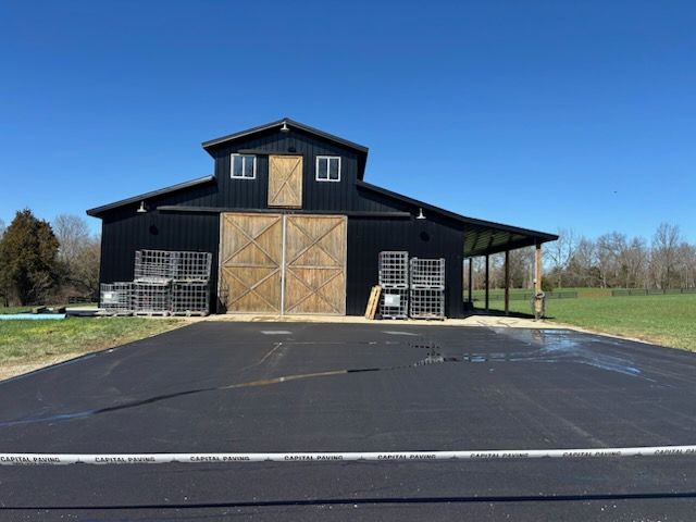 A black barn with large wooden doors, a hayloft, and stacks of metal crates sits behind a freshly paved asphalt driveway.