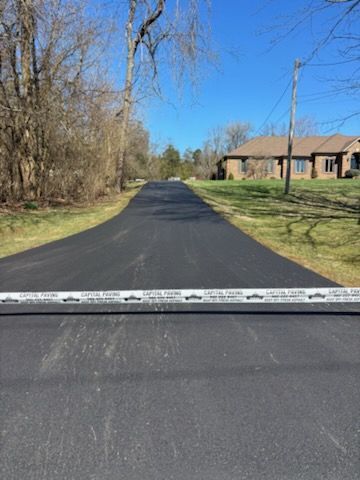 A freshly paved dark asphalt driveway leads to a suburban home, blocked by caution tape on a sunny day.