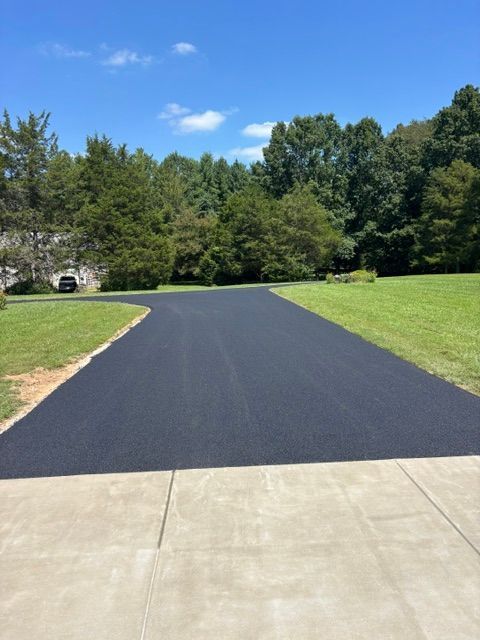 Freshly paved black driveway leading to a wooded yard under a blue sky