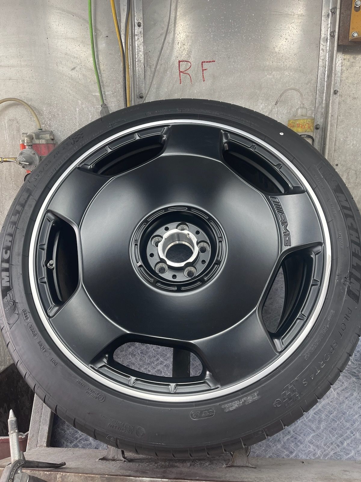 Black car wheel with five-spoke rim and tire, mounted upright in a workshop against a metal wall