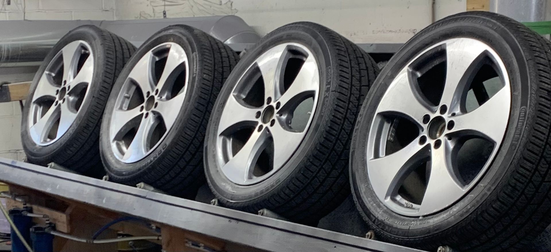 Four silver alloy car wheels with tires lined up on a metal rack outdoors.