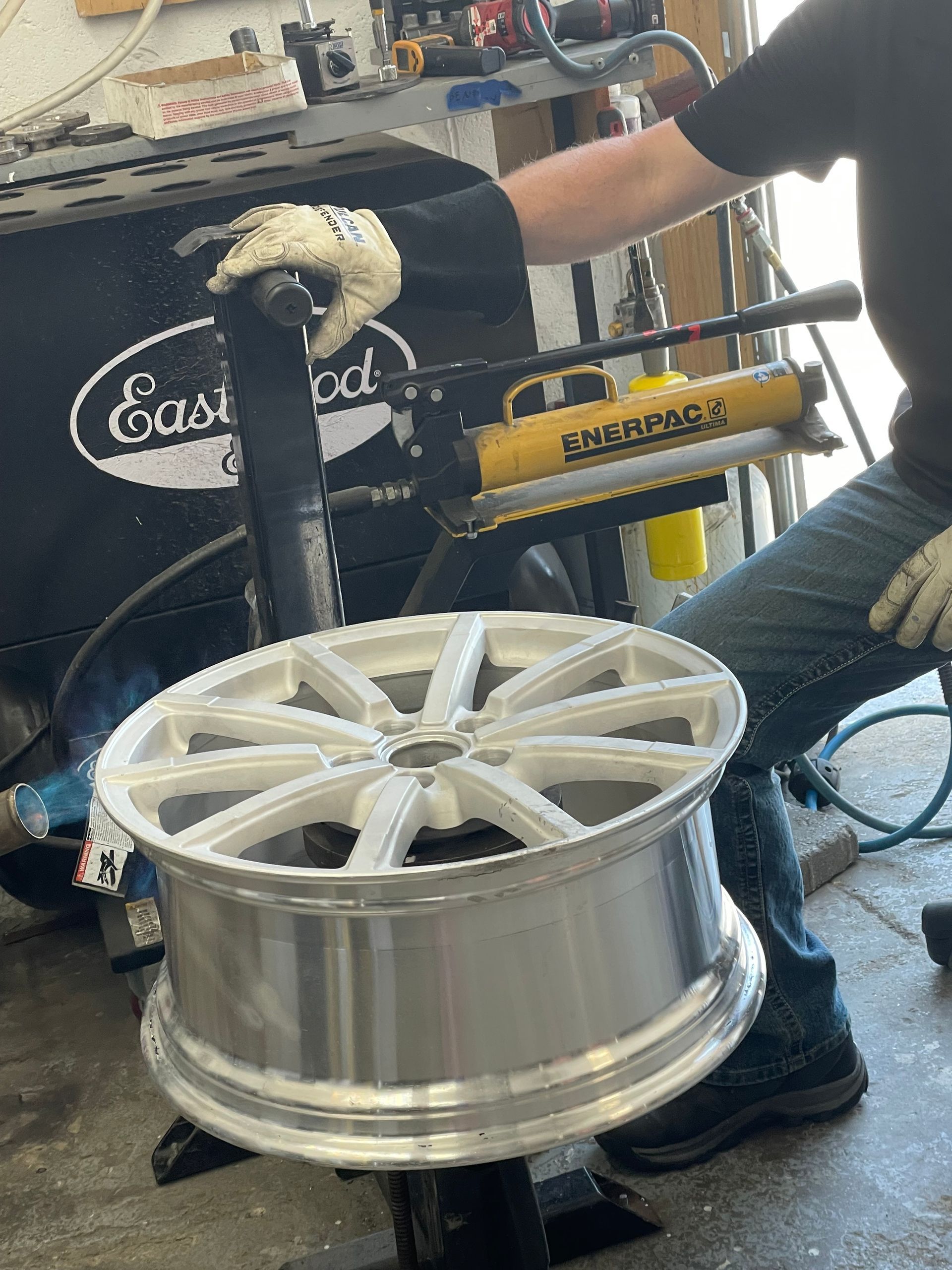 Person holding a large silver car wheel rim in a workshop with tools and equipment in the background