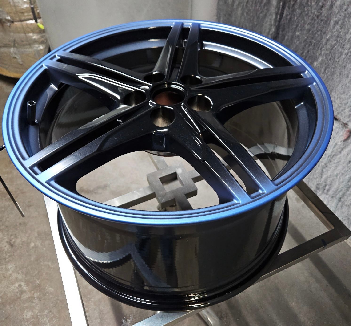Black alloy car wheel rim with a blue rim edge, sitting on a stand in a workshop.