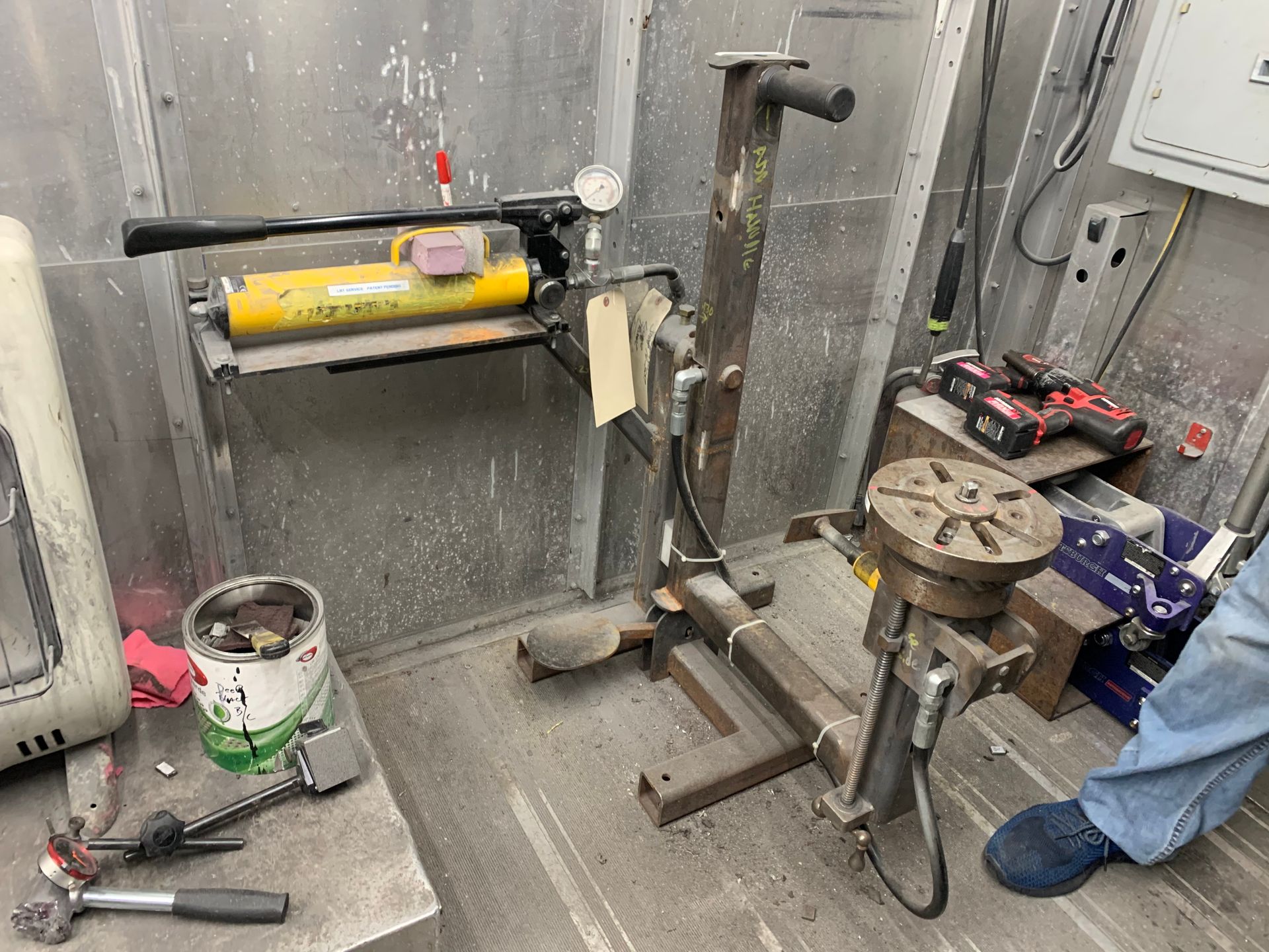 Workshop wall with a yellow caulk gun dispenser and metal pipe assembly beside tools on the floor