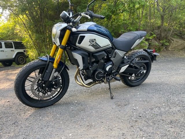 Blue and silver motorcycle parked on a gravel road beside trees, with a white SUV in the background
