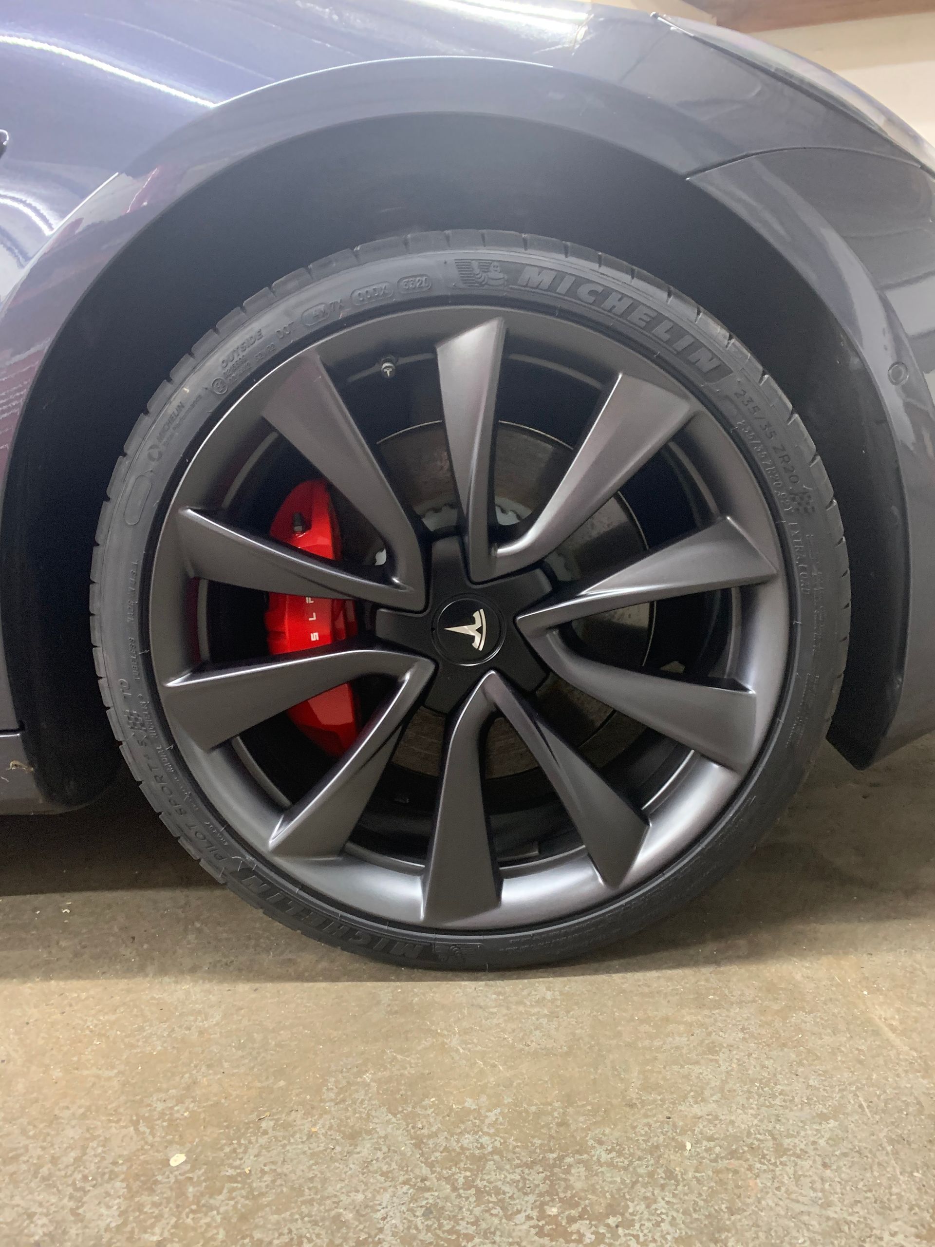 Close-up of a car wheel with black alloy rim, red brake caliper, and tire on a concrete floor.