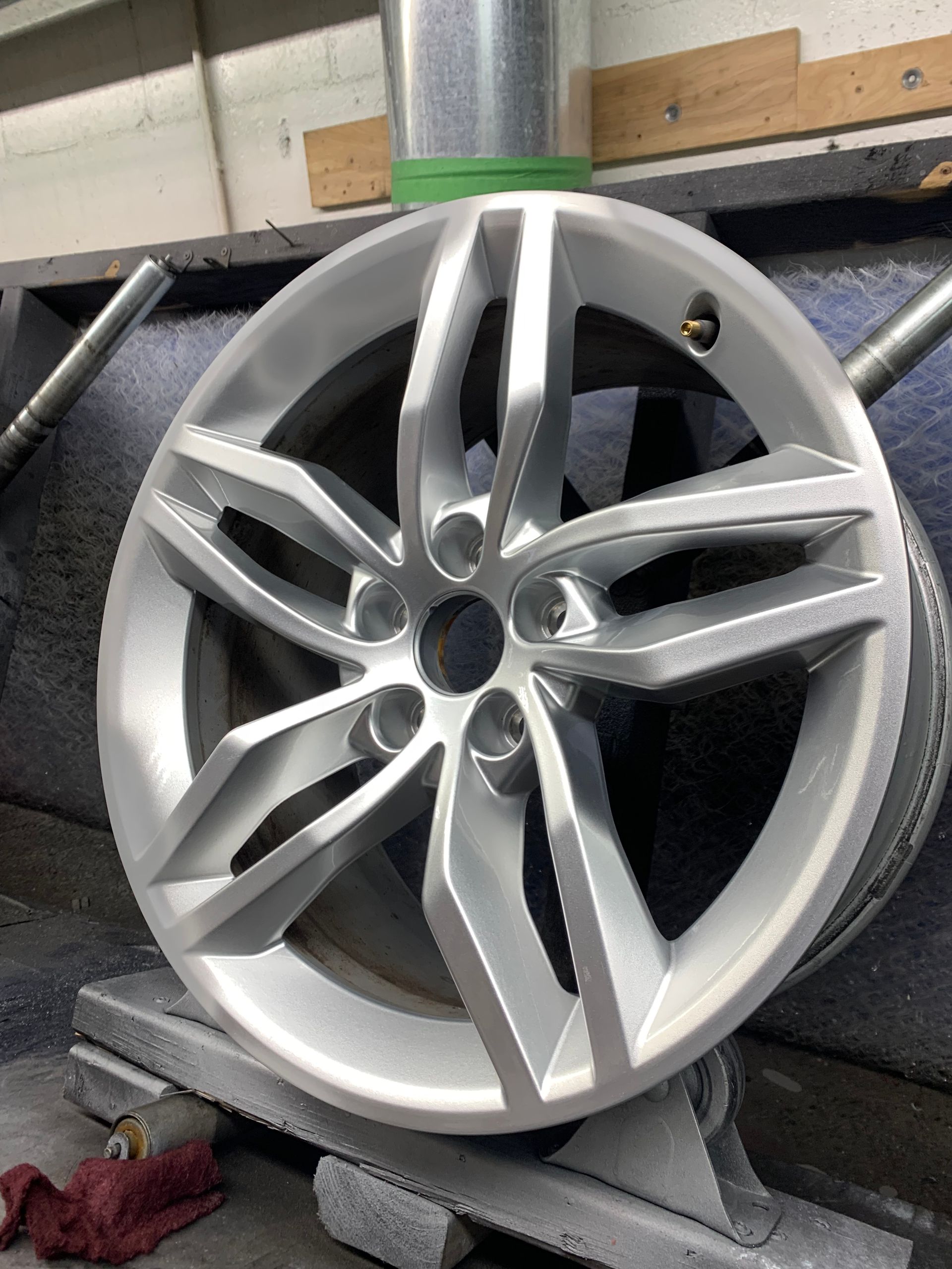 Silver alloy car wheel rim leaning on a foam stand in a workshop, with a tire machine behind it.
