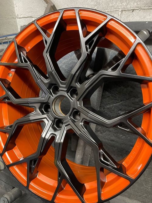 Black multi-spoke car wheel rim with an orange inner barrel on a textured surface