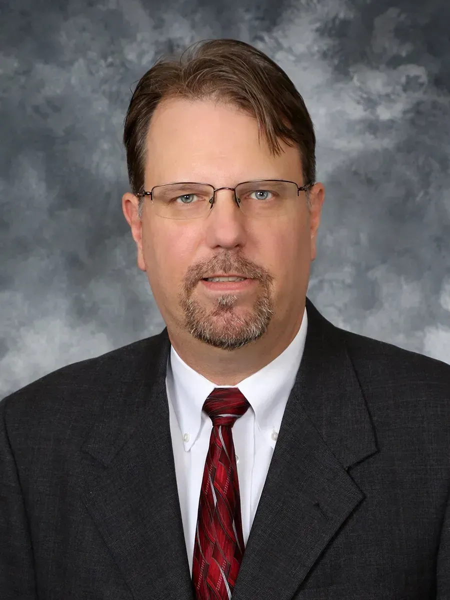 Man with a full beard, hands on head, looking distressed, wearing a gray jacket and cable-knit sweater.