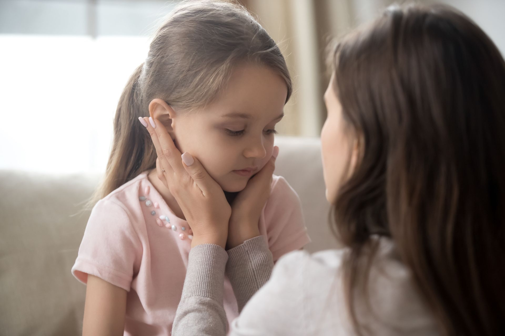 Woman comforting a sad young girl. Woman's hands gently cupping girl's face. Indoors, soft lighting.