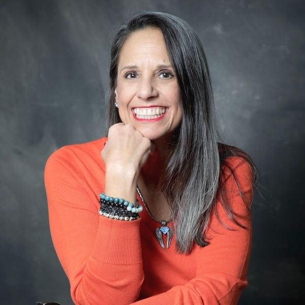 A woman in an orange sweater is smiling with her hand on her chin.