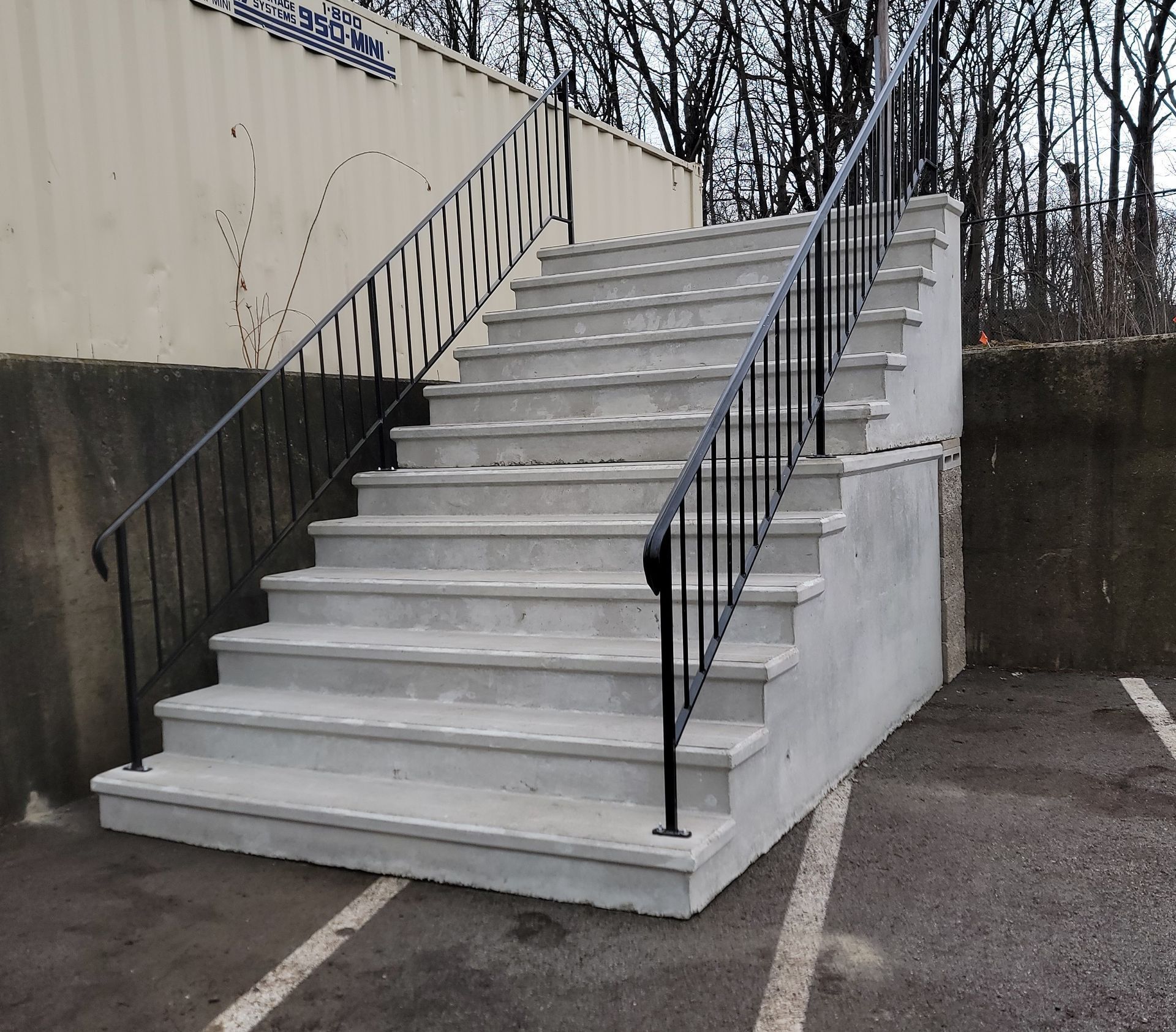 Outdoor concrete staircase with black railings beside a building and retaining wall