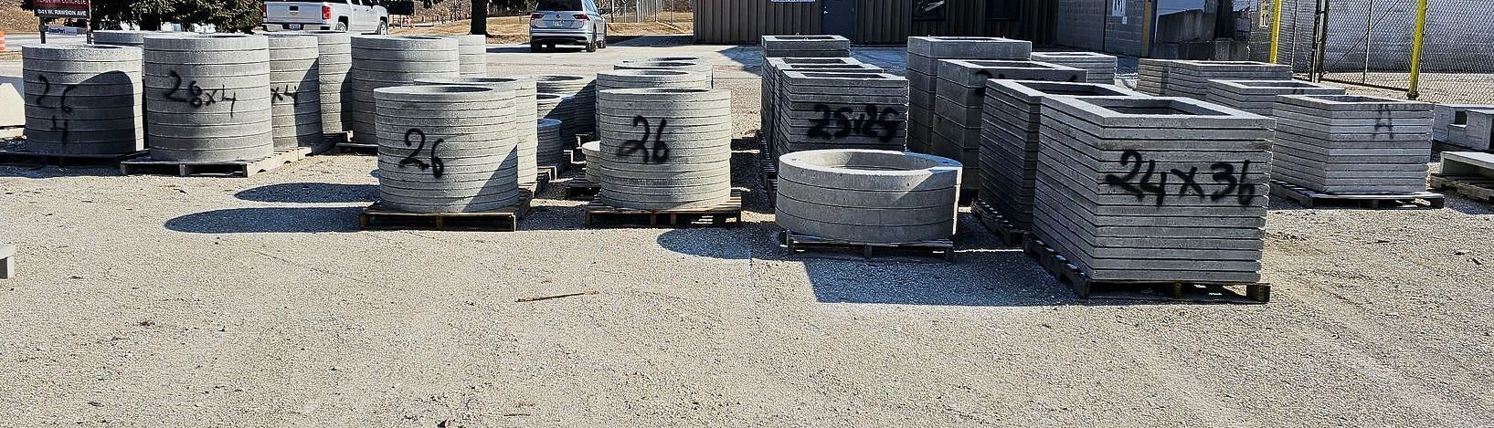 Stacks of gray concrete blocks and pavers arranged on a gravel lot outdoors.